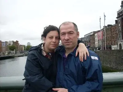 Caterina e Matteo abbracciati su un ponte sul fiume, in una città dal cielo grigio